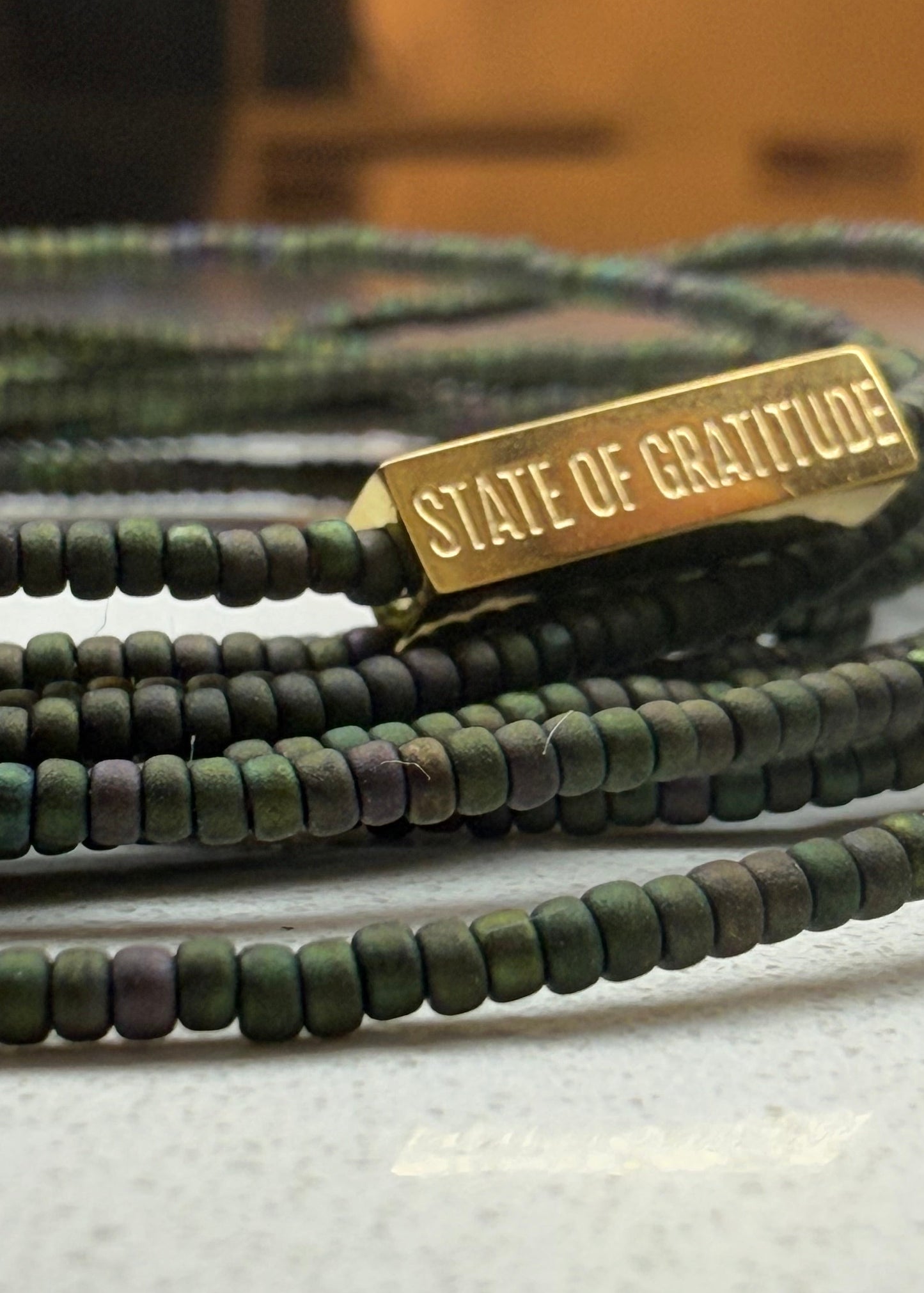 Green beaded bracelet with a gold tag labeled 'State of Gratitude' on a textured surface.