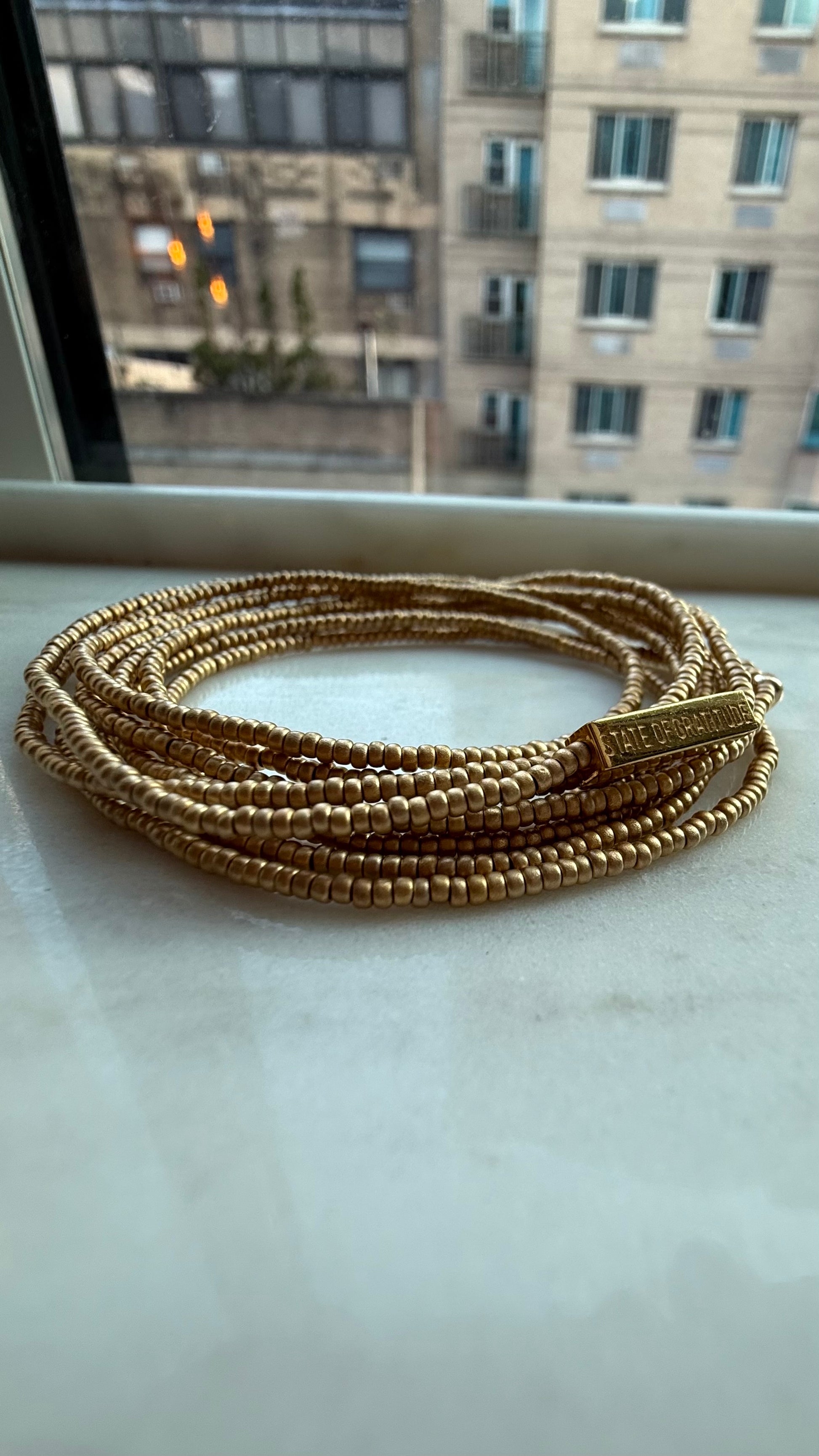 Gold beaded bracelet on a marble surface with a blurred window background